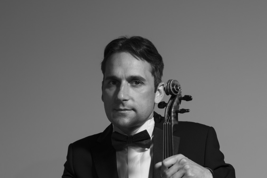 Black-and-white portrait of a person in a tuxedo holding a violin and bow, seated against a plain background.