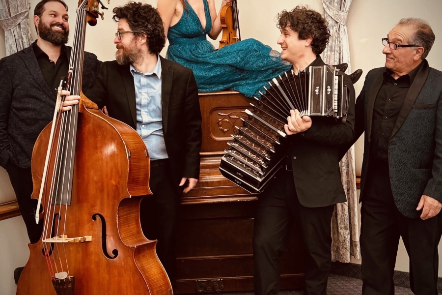 Five musicians indoors with double bass, violin, and accordion, gathered around a piano.
