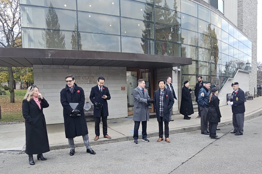 People stand outside a modern building during a remembrance gathering, some wearing coats and poppies along a sidewalk.
