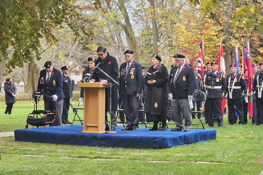 Veterans stand on a small outdoor stage near a podium during a remembrance ceremony, with flags displayed behind them.