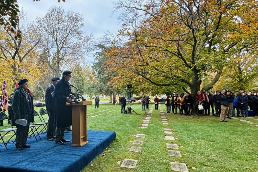 A speaker stands at a podium on an outdoor stage during a remembrance ceremony, with attendees gathered among trees and memorial stones.