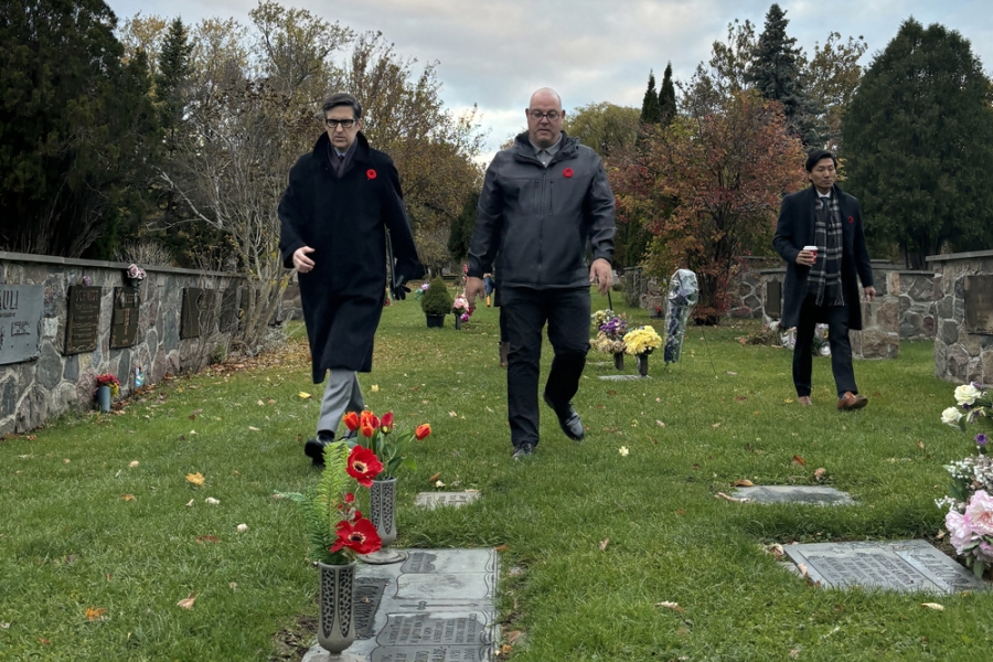 Three people walk through a cemetery during a remembrance visit, with headstones, flowers, and autumn trees visible around them.