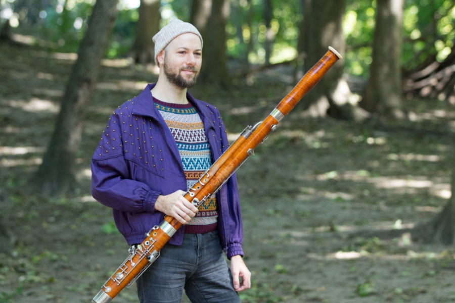 Person standing outdoors in a wooded area holding a large bassoon, wearing a jacket and knit hat.