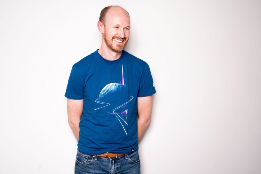 Person standing against a plain background, smiling and wearing a blue graphic T-shirt and jeans.