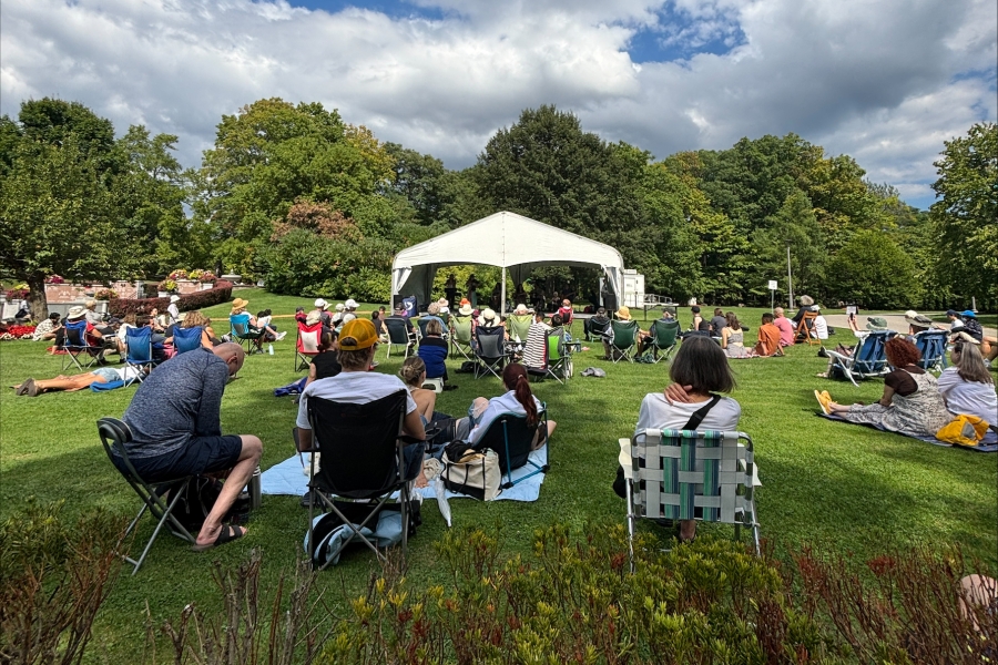 Audience seated on lawn chairs watching a performance under a white tent in a landscaped cemetery, with trees and a partly cloudy sky in the background.
