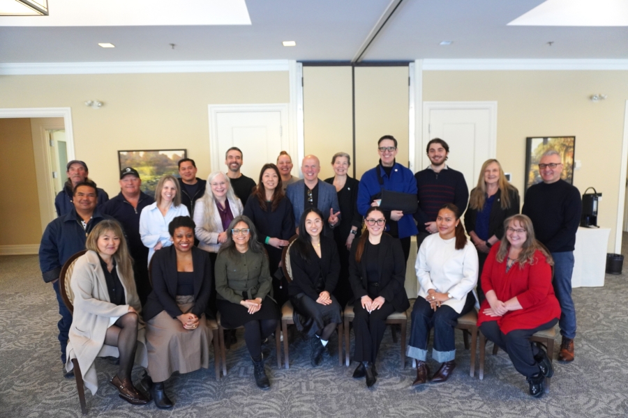 Group photo of about twenty people seated and standing in a conference room with beige walls, framed artwork, patterned carpet, and a folding partition behind them.