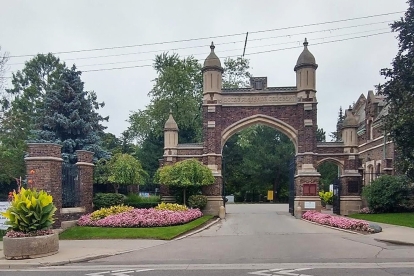 Main entrance to Mount Pleasant Cemetery, featuring a large stone and brick archway with decorative pillars, iron gates, and landscaped flower beds lining the roadway.