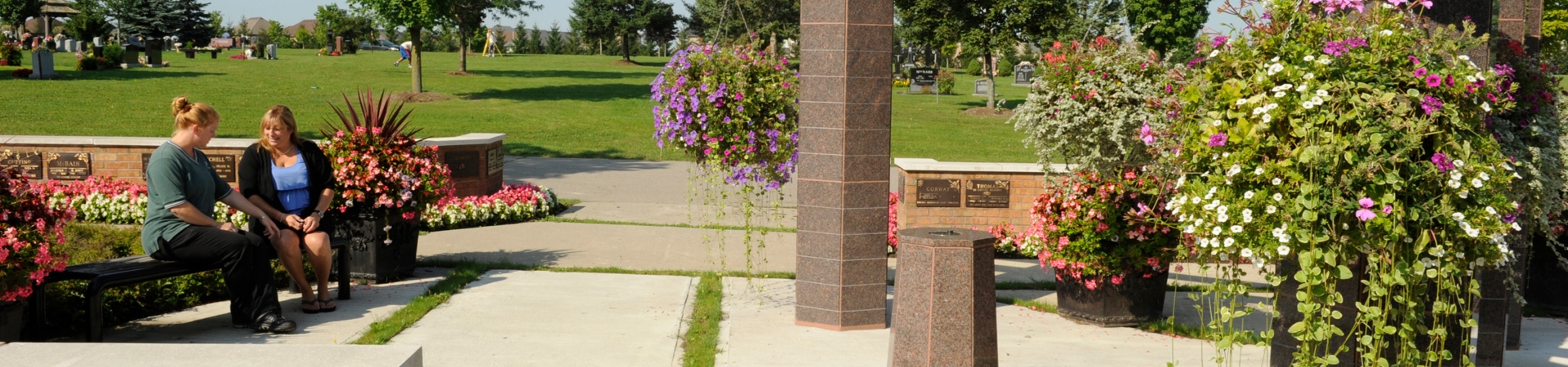 Two people sit on a bench in a landscaped cemetery courtyard with flowers.