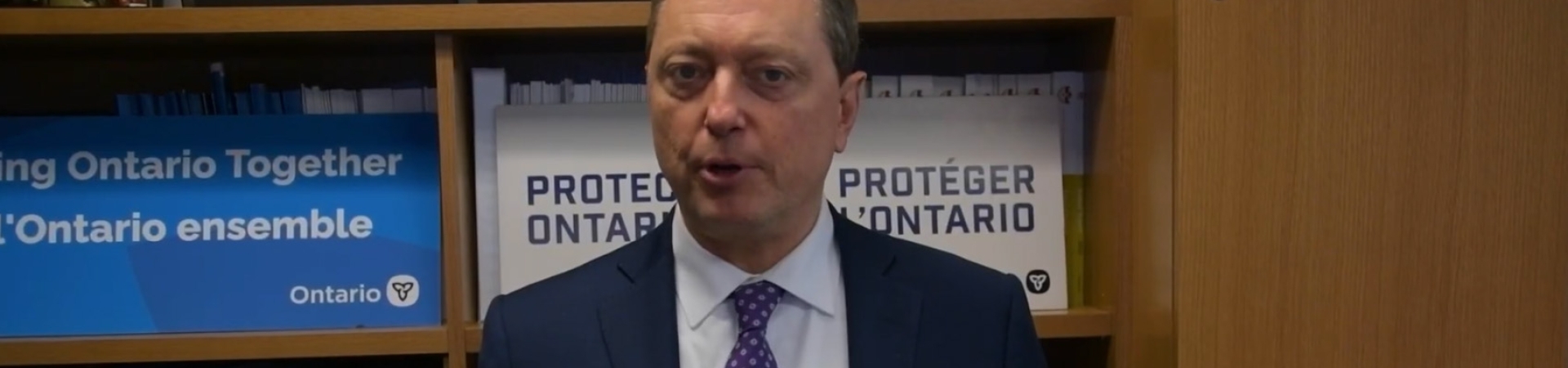 Minister Stephen Crawford speaking in an office, standing in front of shelves with Ontario signage and documents.